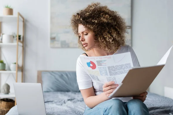 Krullende Vrouw Met Grafieken Mappen Zoek Naar Laptop Bed Thuis — Stockfoto