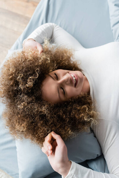 top view of cheerful curly woman with closed eyes stretching on bed at home