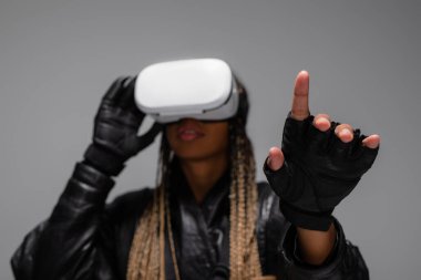 Blurred african american woman in virtual reality headset and gloves pointing with finger isolated on grey  clipart
