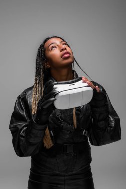 Trendy african american woman holding vr headset and looking up isolated on grey  clipart
