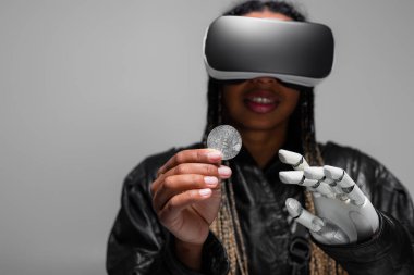 KYIV, UKRAINE - 28 Nisan 2022: VR kulaklıklı ve robot elli bulanık Afrikalı Amerikalı kadın gri üzerinde izole edilmiş bitcoin tutuyor 