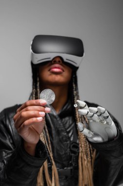 KYIV, UKRAINE - 28 Nisan 2022: Bulanık Afro-Amerikan kadın vr kulaklıklı robot eli Bitcoin gri üzerinde izole edilmiş 