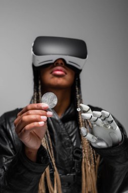 KYIV, UKRAINE - APRIL 28, 2022: Blurred african american woman in vr headset with robotic hand holding bitcoin isolated on grey  clipart