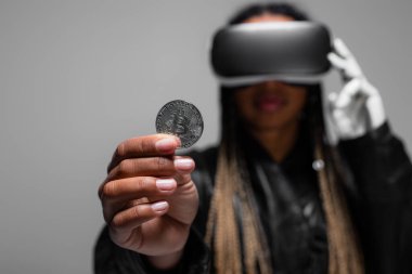 KYIV, UKRAINE - 28 Nisan 2022: Bulanık Afro-Amerikan kadın VR kulaklıklı gri üzerinde bitcoin tutan 