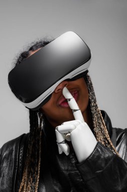 VR kulaklıklı ve robot elli Afrikalı Amerikalı kadın. Gri renkte izole edilmiş bir jest gösteriyor. 