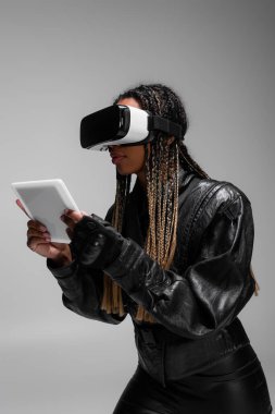 Gri üzerine izole edilmiş dijital tablet kullanan deri ceketli ve video kulaklı Afrikalı Amerikalı kadın. 