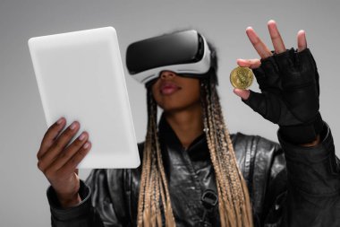KYIV, UKRAINE - 28 Nisan 2022: Gri üzerinde bitcoin ve dijital tablet taşıyan VR kulaklıklı Afro-Amerikan kadın 