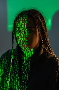 Gri arkaplandaki kameraya bakan bilgisayar kodu projeksiyonlu Afrikalı Amerikalı kadın. 