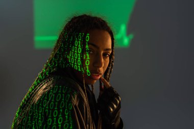 Gri arka planda bilgisayar kodu yansıması olan deri ceketli Afrikalı Amerikalı kadın. 