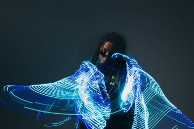African american woman in smart glasses looking at camera near abstraction neon lights on grey background  clipart