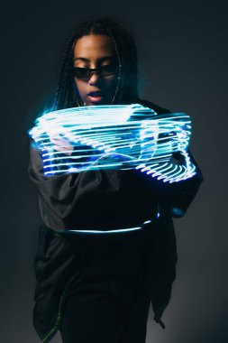 Beyaz gözlüklü şık Afro-Amerikalı kadın gri arka plandaki neon ışıklara bakıyor. 