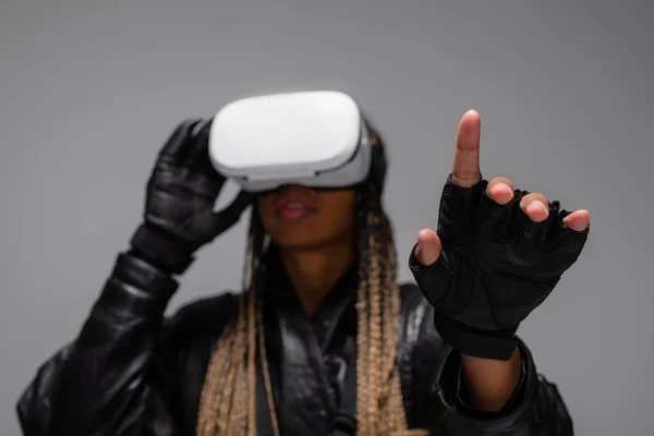 Blurred african american woman in virtual reality headset and gloves pointing with finger isolated on grey