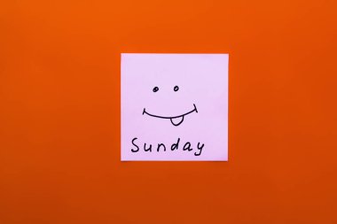 top view of paper with sunday lettering and cheerful smiley with sticking out tongue on orange background clipart
