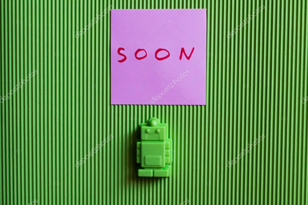 Top view of toy robot near purple paper with soon lettering on green textured background