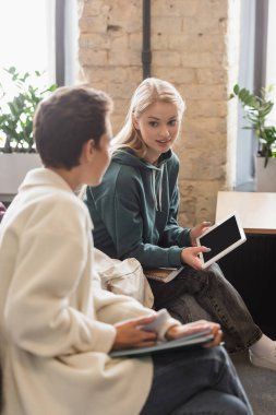 Dijital tabletli gülümseyen kadın sınıfta bulanık arkadaşıyla konuşuyor.