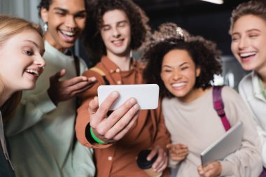 Genç adam cep telefonuyla selfie çekiyor. Farklı ırklardan gelen öğrencilerle.