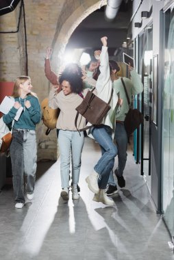 Üniversite koridorunda yürüyüp neşeli hareketler yapan çok ırklı arkadaşlar.