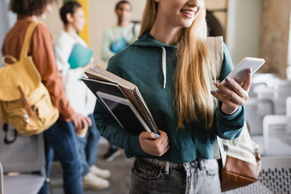 smiling woman with copy books and digital tablet using smartphone near blurred classmates