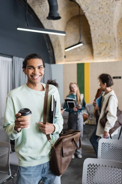 cheerful african american man with leather bag and coffee to go near students in blurred classroom