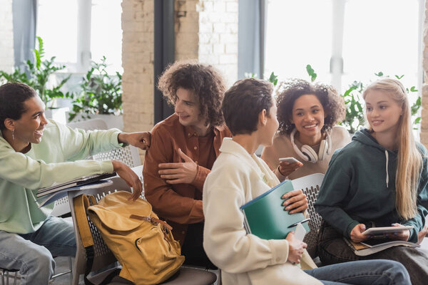 cheerful multicultural students with copybooks and gadgets talking while sitting in auditorium