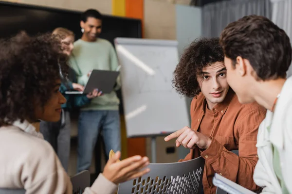 curly student pointing with finger during discussion with interracial classmates in auditorium