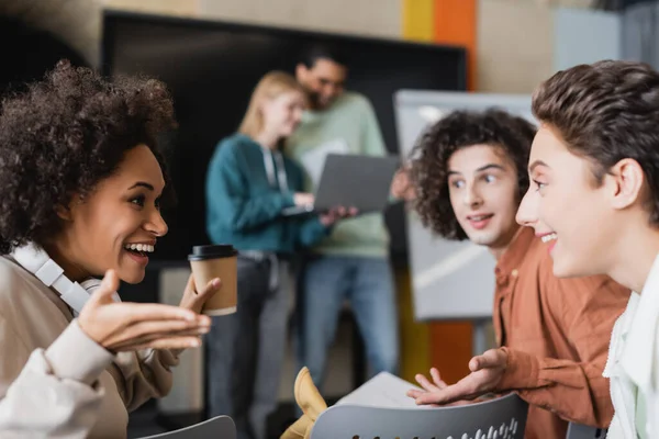 excited african american student with paper cup gesturing during discussion with classmates