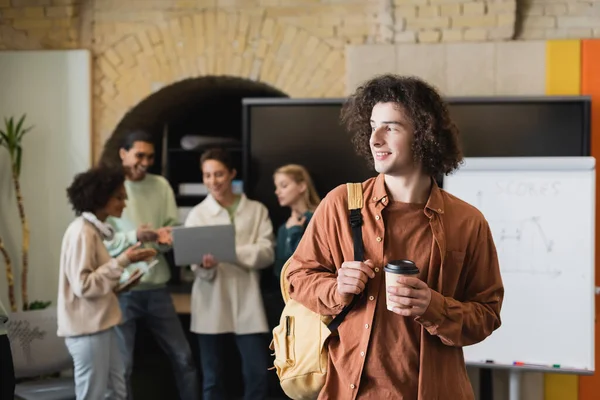 curly man with backpack and coffee to go looking away while blurred interracial classmates talking near laptop