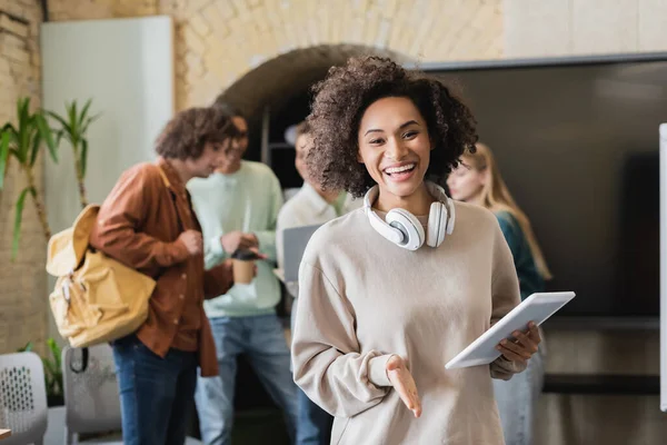 cheerful african american woman with headphones and digital tablet looking at camera near blurred students