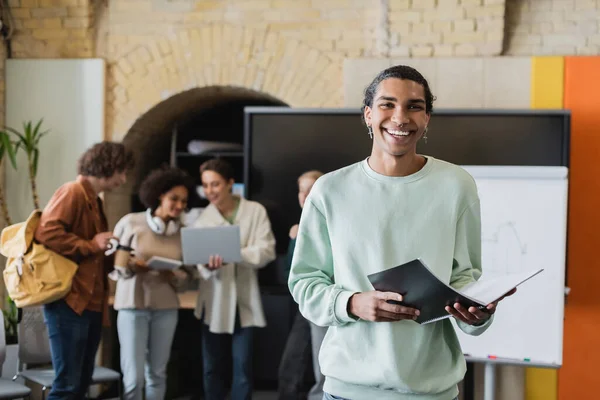 african american student with notebook smiling at camera near blurred interracial classmates with gadgets