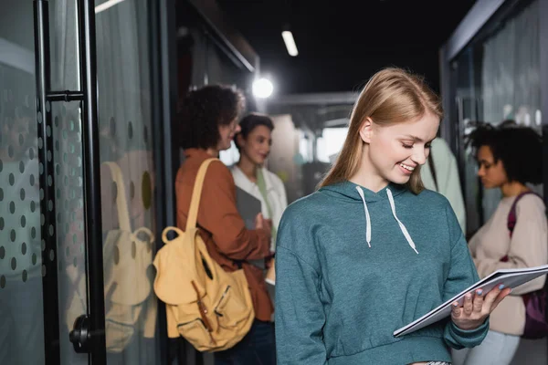 Smiling Woman Looking Copybook Multicultural Classmates Blurred Hallway — Stock Photo, Image