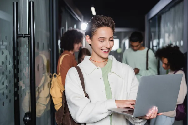 Radosna Kobieta Korzystająca Laptopa Stojąc Pobliżu Międzyrasowych Studentów Zamglonym Korytarzu — Zdjęcie stockowe