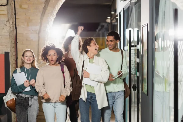 excited woman showing win gesture near interracial friends in university corridor