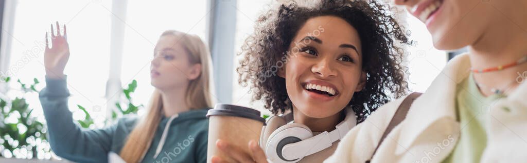 happy african american woman with coffee and headphones looking at blurred friend, banner