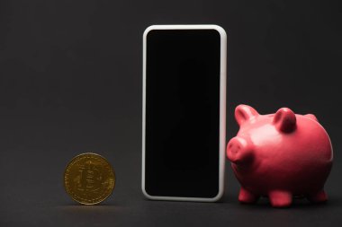 KYIV, UKRAINE - 26 Nisan 2022: Domuzcuk kumbarası yakınındaki akıllı telefon ve siyah arka plandaki bitcoin görüntüsünü kapat