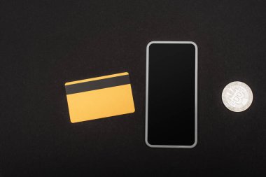 KYIV, UKRAINE - 26 Nisan 2022: Cep telefonu yakınındaki kredi kartının üst görüntüsü ve siyah arka plandaki bitcoin