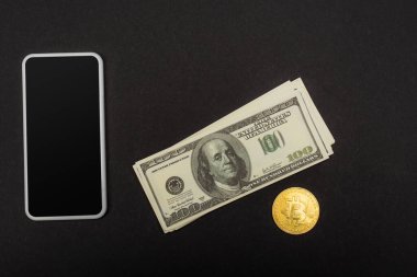 KYIV, UKRAINE - 26 Nisan 2022: Dolarların yanında boş ekran ve siyah arka planda altın bitcoin ile akıllı telefonun üst görüntüsü