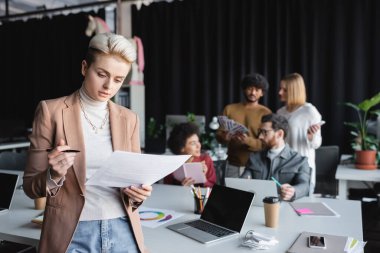Belgeleri ve kalemleri bulanık arkaplanda cihazları ve ırklar arası meslektaşları olan bir tasarımcı.