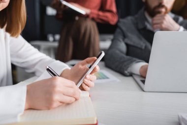 Reklam ajansında not defterine akıllı telefon yazan kadının kısmi görüntüsü