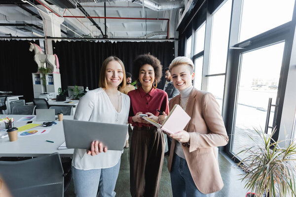 cheerful multiethnic women with notebook and gadgets looking at camera in ad agency