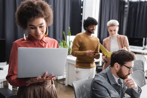 positive african american woman looking at laptop near multiethnic colleagues working in ad agency