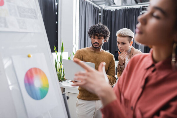 blurred african american woman pointing at flip chart near thoughtful interracial colleagues with laptop