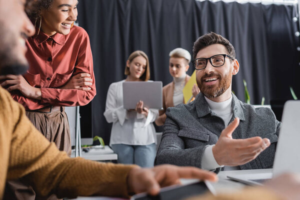 happy advertising manager in eyeglasses pointing at blurred laptop near interracial colleagues