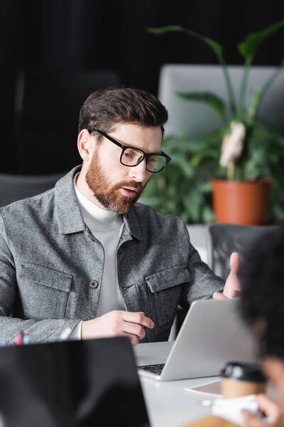 bearded designer in eyeglasses working near laptop in ad agency near blurred colleague