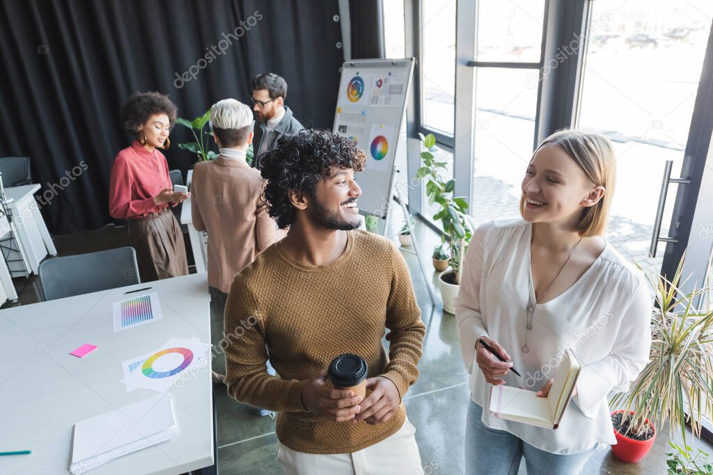 cheerful interracial advertising agents with paper cup and notebook looking at each other in office