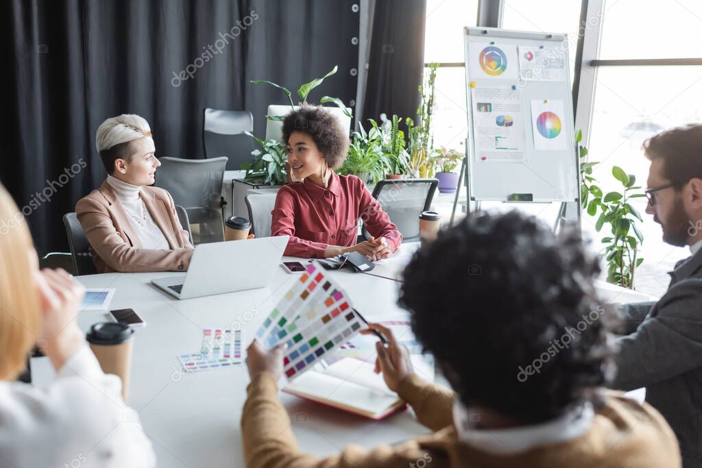 smiling interracial women talking to each other during meeting with colleagues in advertising agency