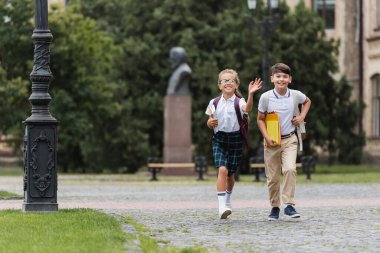 Elinde defterlerle gülümseyen liseli kız Asyalı arkadaşının yanında el sallıyor. 