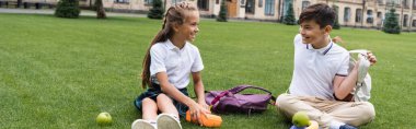 Smiling schoolgirl holding lunchbox near asian friend with backpack on grass in park, banner  clipart