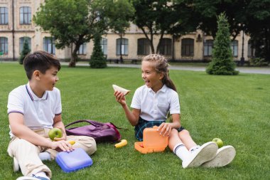Excited schoolgirl holding sandwich near lunchbox and asian friend with apple on lawn  clipart