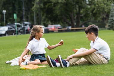 Side view of schoolchild holding sandwich near asian classmate on grass in park  clipart