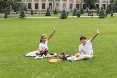 Parktaki çimlerin üzerinde sandviç tutan ve el sallayan neşeli ırklar arası okul çocukları. 